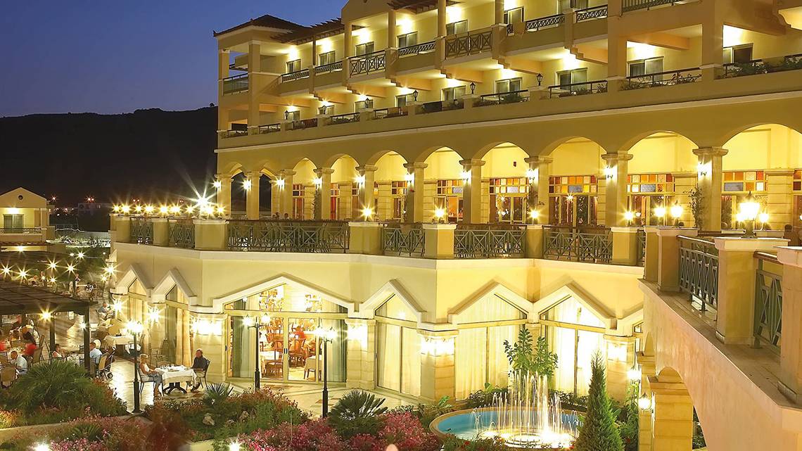 Außenansicht des Lindos Princess Beach Hotels bei Nacht mit beleuchteten Balkonen und Brunnen.