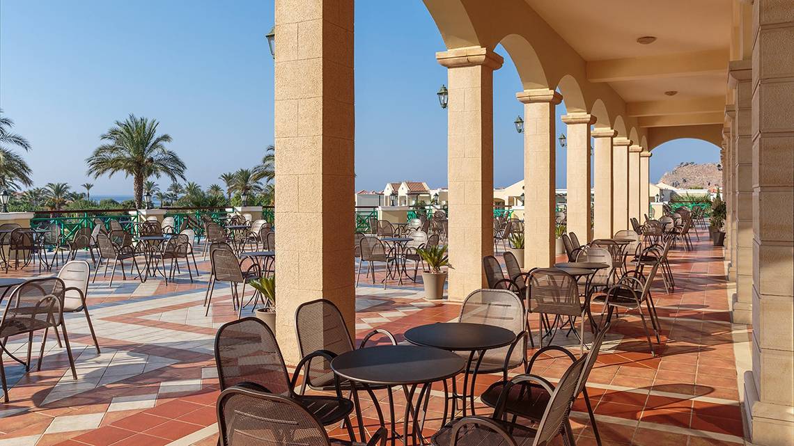 Außenbereich des Lindos Princess Beach Hotels mit vielen Tischen und Stühlen unter Arkaden.