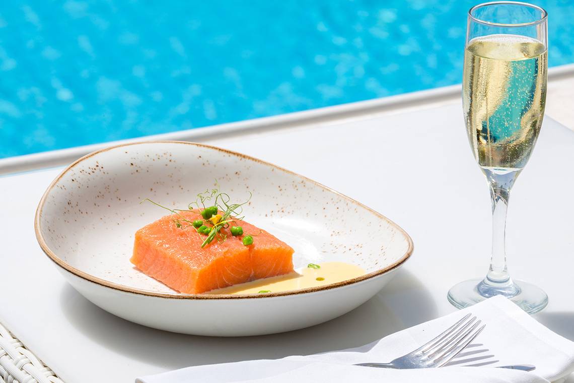 Ein Teller mit Lachs in einer Porzellanschale, neben einem Glas Champagner, vor einem Pool.
