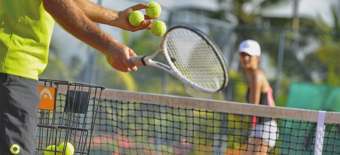 Tennisspieler hält Tennisschläger und Tennisbälle auf einem Tennisplatz mit Netz im Hintergrund.