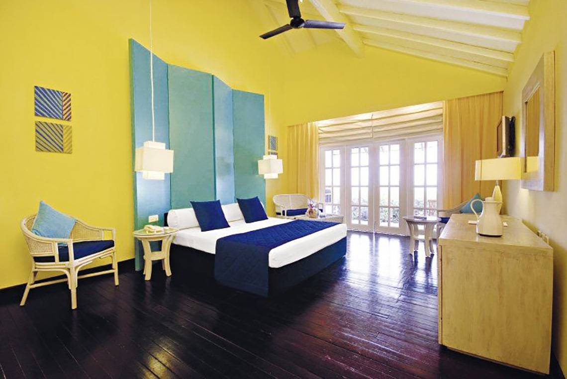 Hotelzimmer im Adaaran Select Meedhupparu mit gelben Wänden, blauer Bettwäsche und einem offenen Fenster.