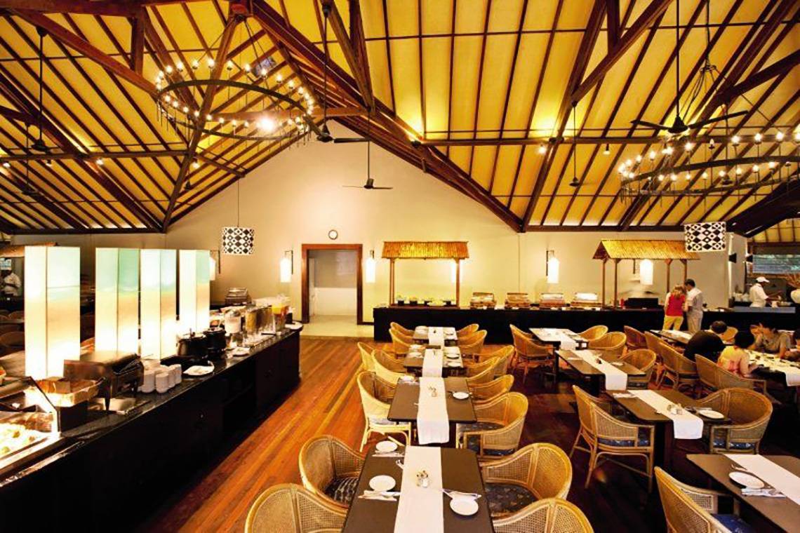Innenansicht des Restaurants im Adaaran Select Meedhupparu Hotel mit Holzboden, Tischdecken und gedimmten Kronleuchtern.