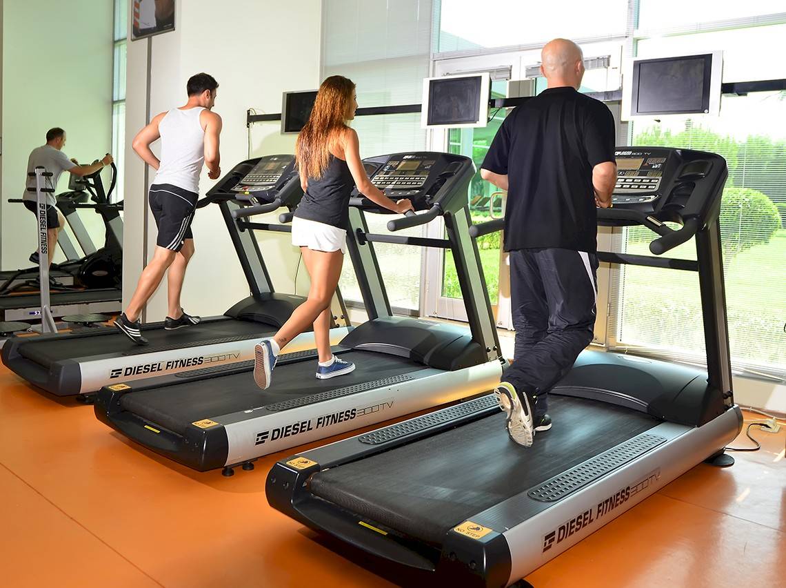 Fitnessstudio mit mehreren Personen, die auf Laufbändern trainieren, darunter ein Mann in einem weißen Tanktop.