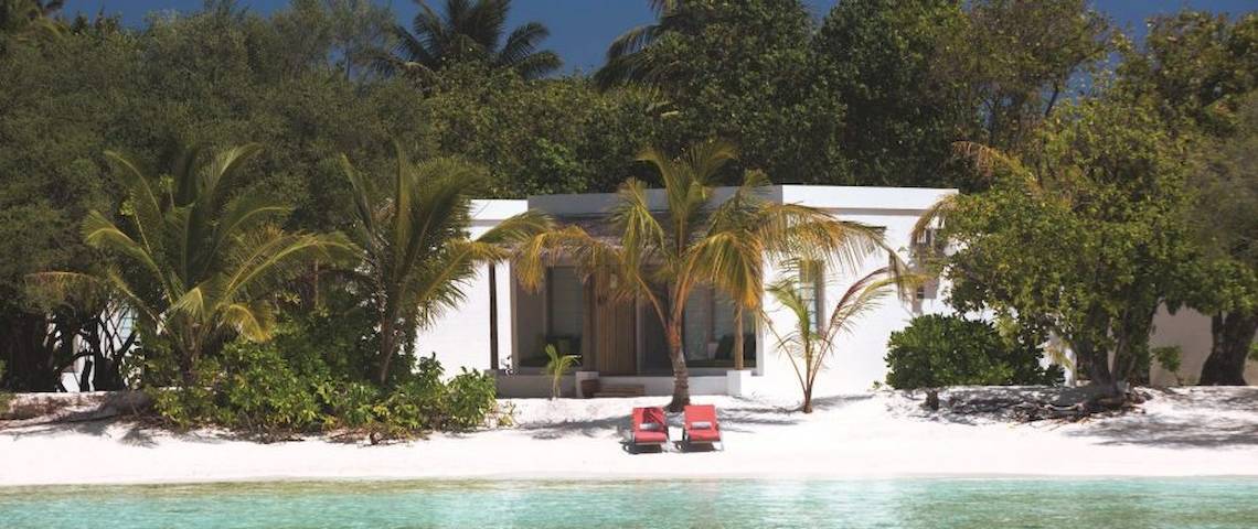 Weißes Hotel OBLU NATURE Helengeli am Strand mit zwei roten Sonnenliegen und Palmen