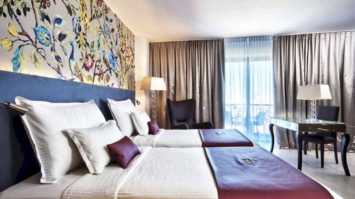 Doppelbett-Zimmer im AX The Palace Hotel mit kunstvollen Wandgemälden, zwei Betten, Tisch, Lampen und Stühlen.