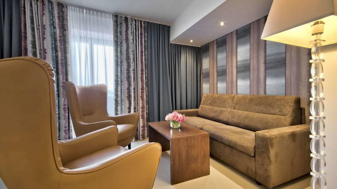 Innenansicht des AX The Palace Hotels mit einem Wohnzimmer, Sofa, zwei Ledersesseln, Tisch, Blumenvase und einer Stehlampe.