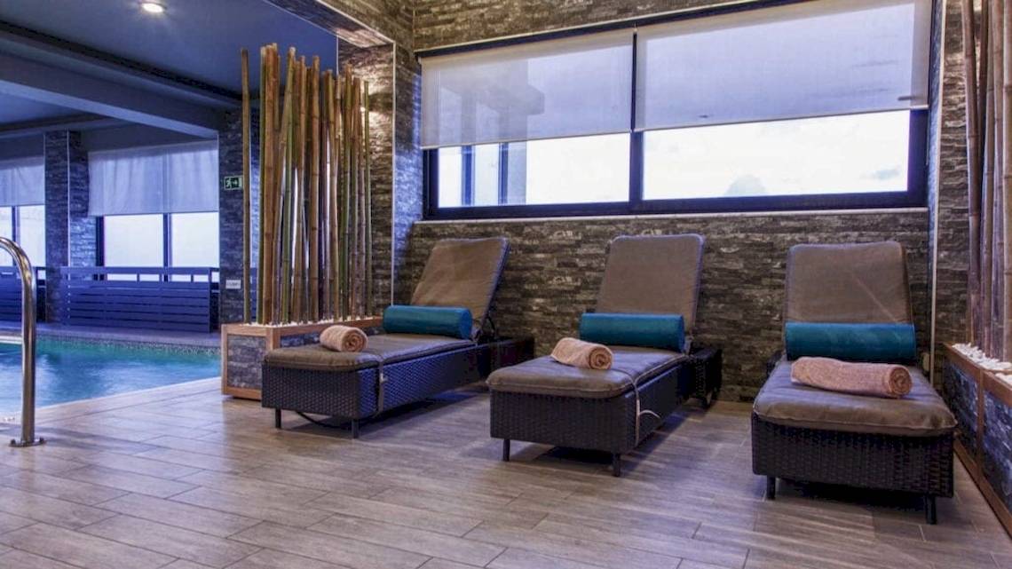 Spa-Lounge-Bereich im AX The Palace Hotel mit drei Liegen, die an einer Wand mit Steinmuster und Glasfenstern ausgerichtet sind.