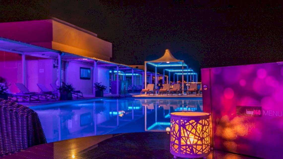 Nachtansicht des Swimmingpools des AX The Palace Hotels mit farbigen Lichtern und Loungestühlen.