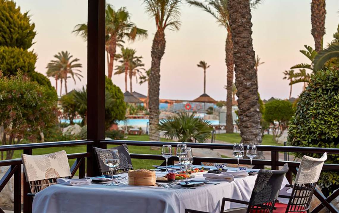 Terrasse mit Tisch und Blick auf Palmen und eine Poollandschaft im Atlantica Aegean Blue Resort.