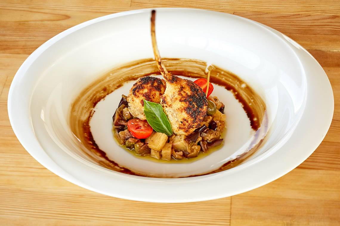 Ein Gericht in einem weißen Teller auf einem Holztisch mit gebratenem Fleisch, Gemüse und Sauce.
