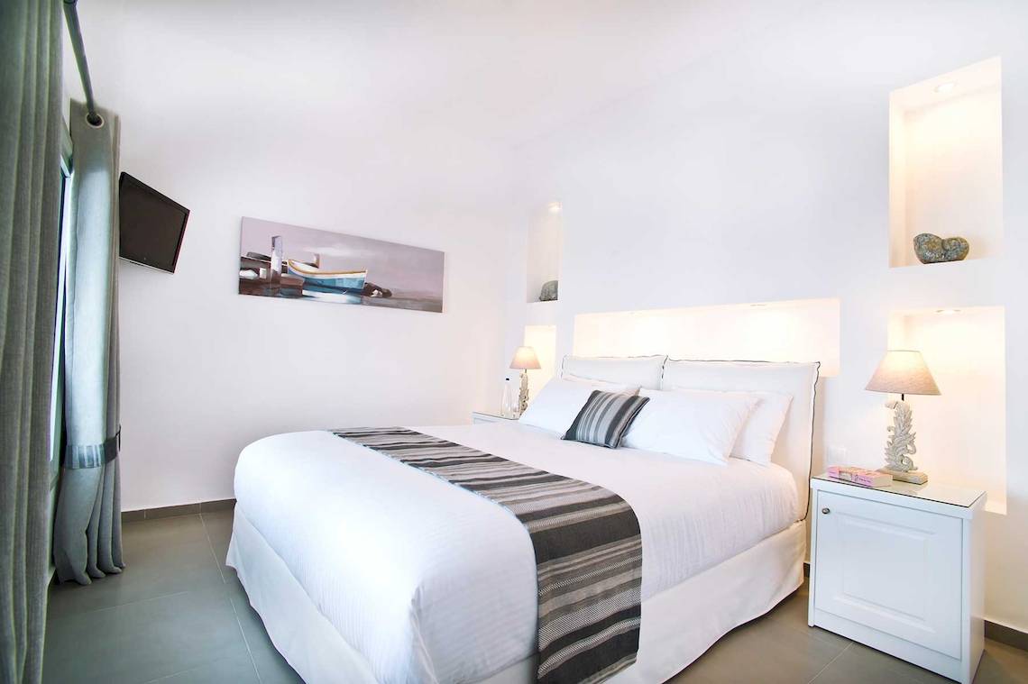 Schlafzimmer im Hotel Agali Houses mit weißem Bett, Kissen, Lampen und einem Fernseher an der Wand.