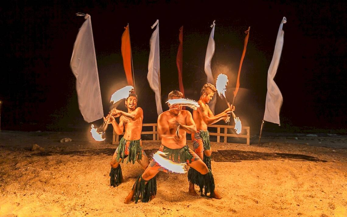 Drei Männer führen eine Feuershow am Strand vor, während sie Fackeln halten und Fahnen im Hintergrund stehen.