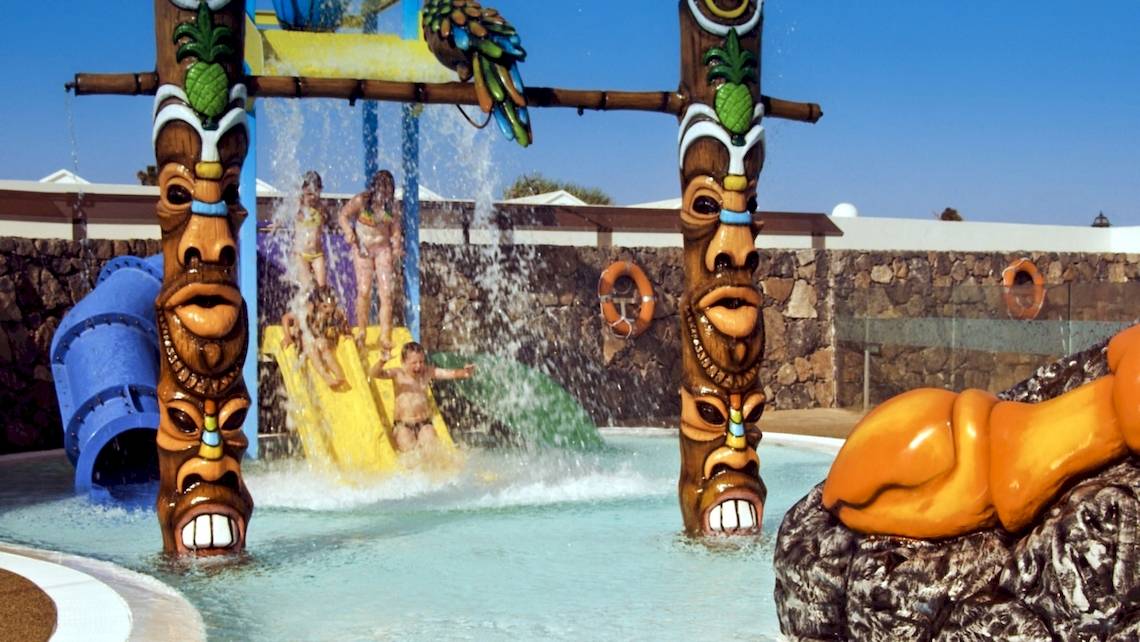 Kinder spielen auf dem Wasserspielplatz des HD Beach Resorts mit bunten Wasserrutschen und Tiki-Figuren.