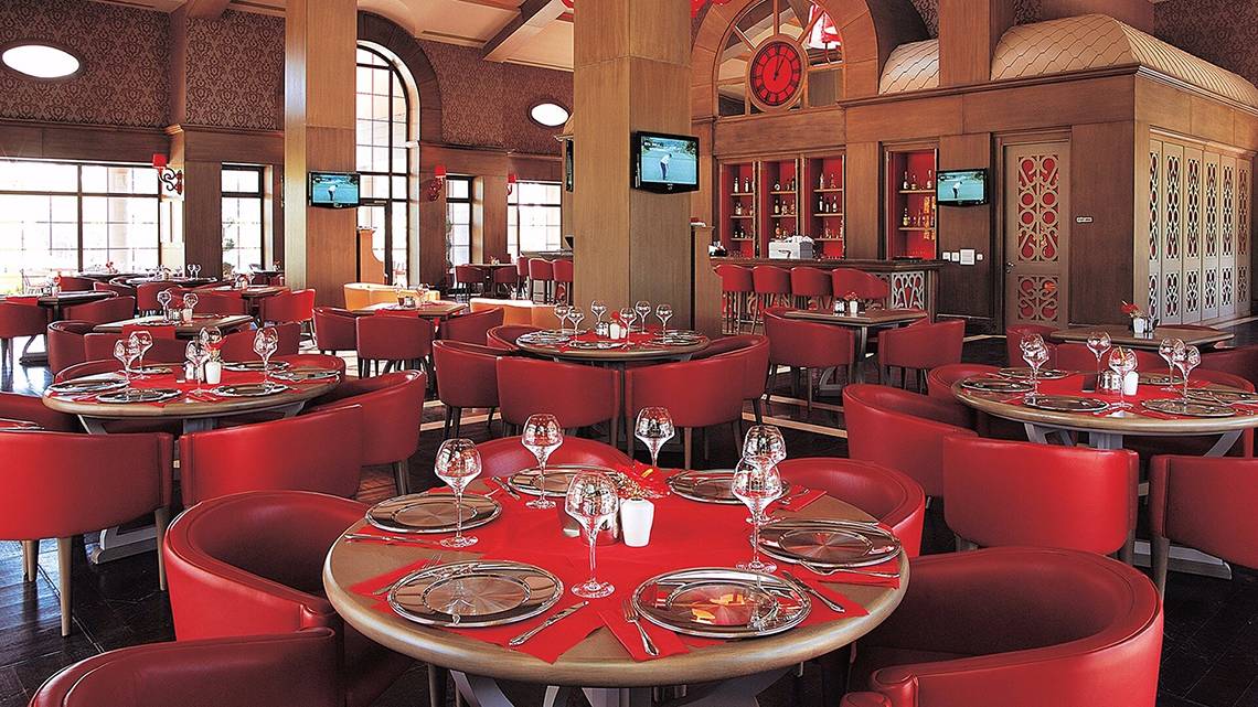 Restaurant im Cornelia Diamond Golf Resort & Spa mit roten Stühlen und Tischen, Fernsehgeräten und Weinflaschen.