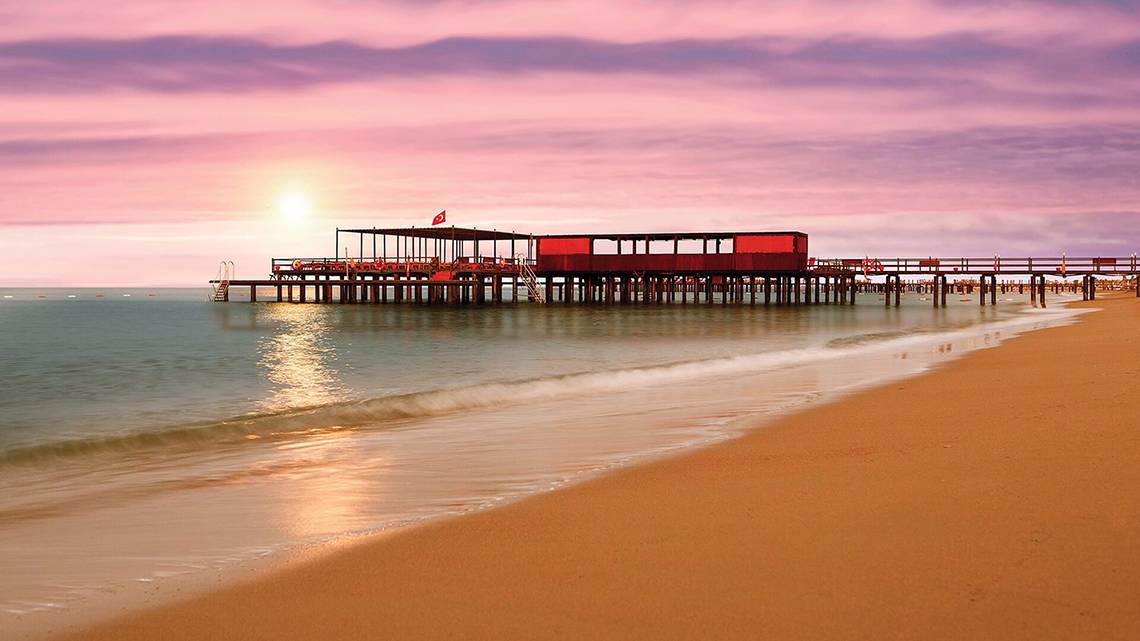 Sonnenuntergang am Pier des Cornelia Diamond Golf Resort & Spa mit rotem Gebäude und Flagge.