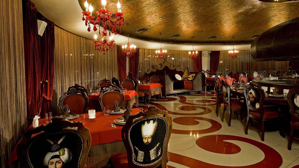 Restaurant-Innenbereich im Cornelia Diamond Golf Resort & Spa mit roten Tischdecken und goldenen Kronleuchtern.