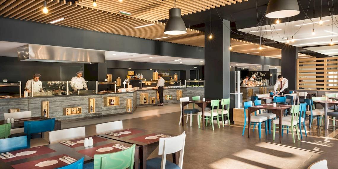 Innenansicht des Restaurants im Remisens Hotel Epidaurus mit Küchenbereich, Tischen und Stühlen.