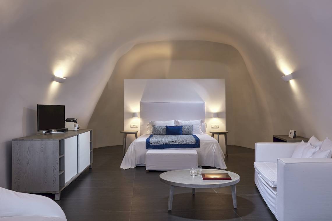 Ein modernes Schlafzimmer im Katikies Chromata Santorini Hotel mit weißem Bett, Nachttischen und einem Fernseher.