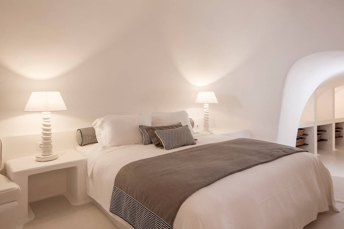 Ein geräumiges Hotelzimmer im Katikies Chromata Santorini mit weißen Wänden, zwei Lampen, einem Bett und einem weißen Nachttisch.