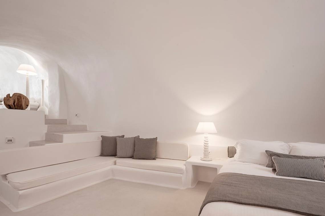 Weißes Zimmer im Katikies Chromata Hotel auf Santorini mit Bett, Sofa und Lampen.