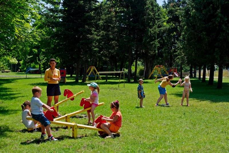 Kinder spielen im Park mit Rutschbahnen und Schaukeln unter Aufsicht von Erwachsenen.