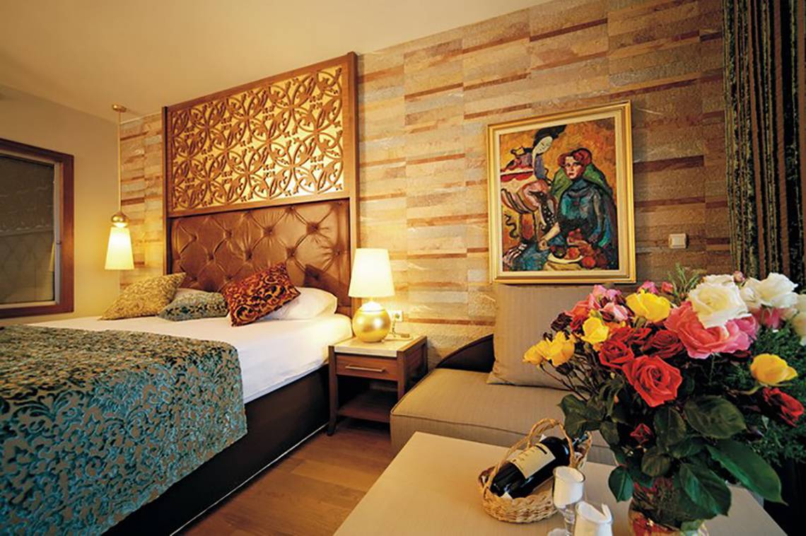 Innenansicht eines Zimmers im Melas Lara Hotel mit Bett, Nachttisch, Gemälde und Blumen.