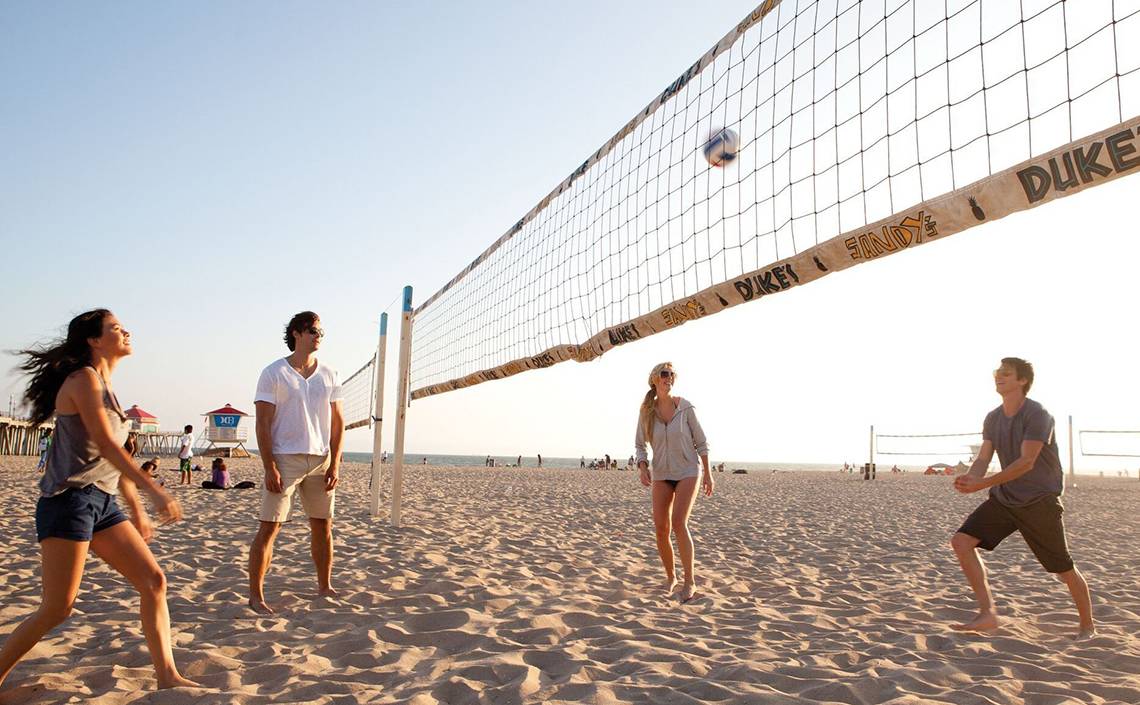 Vier Personen spielen Volleyball am Strand mit einem Netz, das Duke und Sandy & Duke beschriftet ist.
