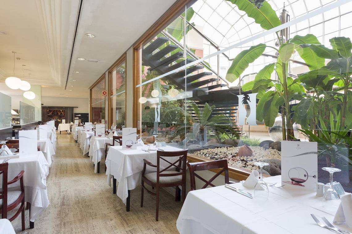 Innenansicht des Restaurants im Hipotels La Geria mit gedeckten Tischen und Stühlen vor großen Glasfenstern.