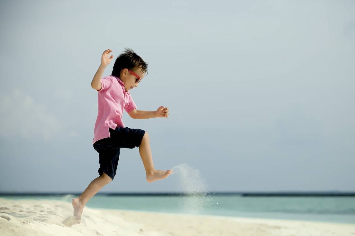 Kleiner Junge springt auf dem weißen Sandstrand und verspritzt Sand