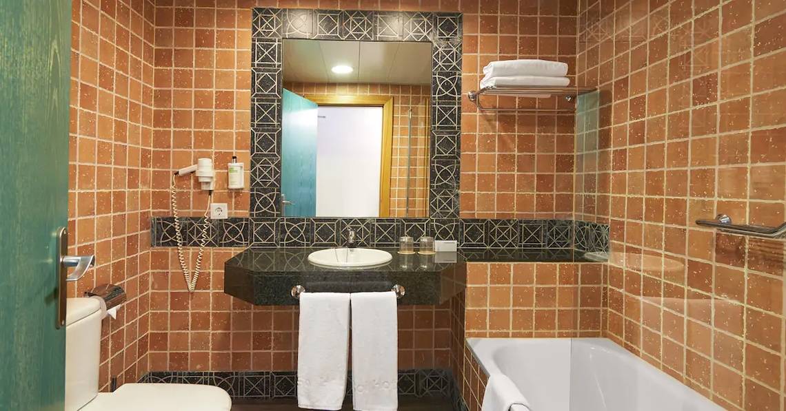 Badezimmer im Sol Barbados Hotel mit braunen Fliesen, einem Waschbecken, einem Spiegel und einer Badewanne.