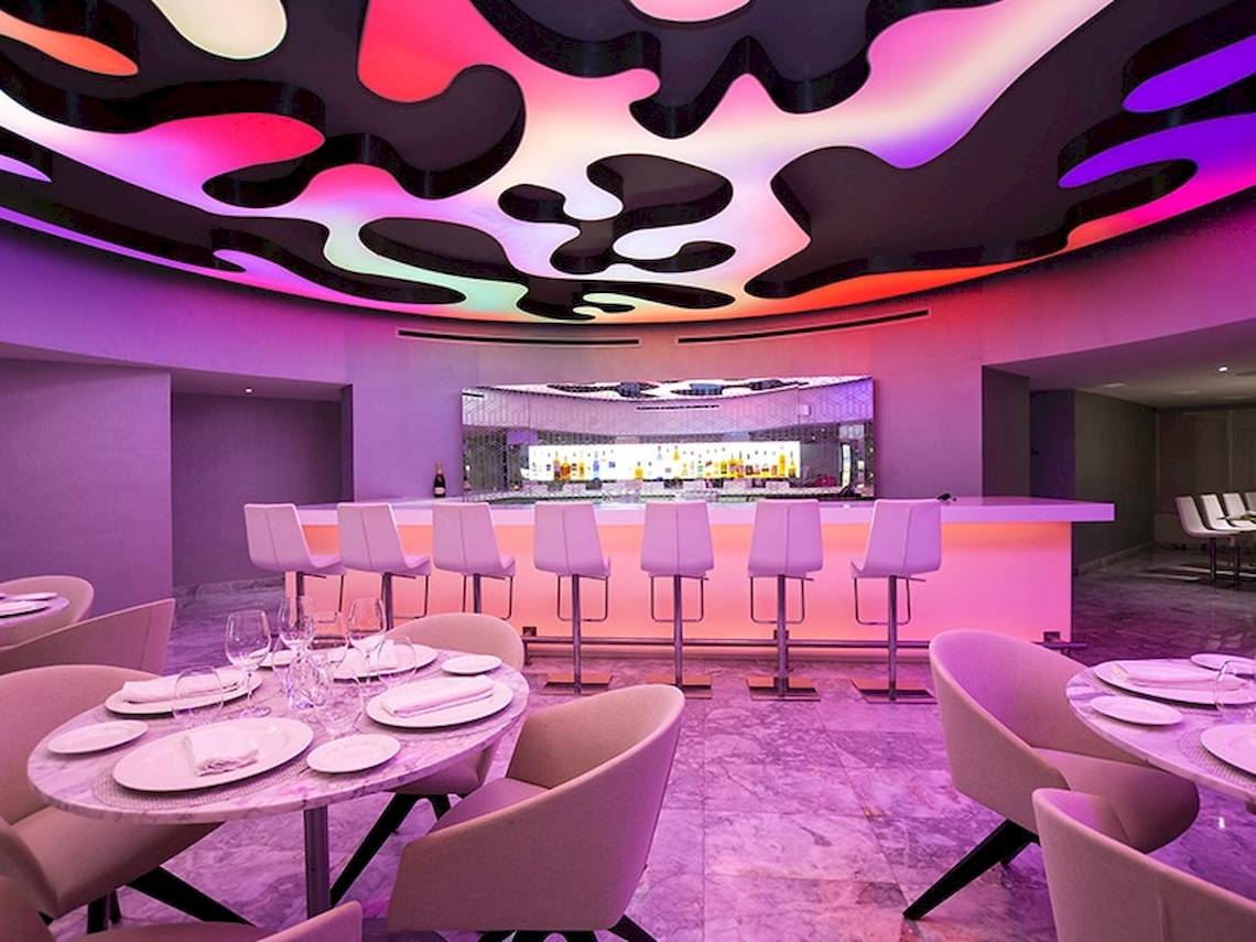 Hotel-Bar-Lounge-Bereich mit Tisch, Barhockern und violett-pinker Beleuchtung.