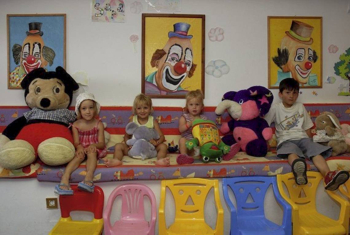 Vier Kinder sitzen auf bunten Stühlen neben großen Plüschtieren und drei Clown-Gemälden an der Wand.