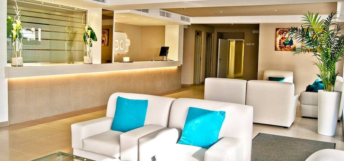 Empfangsbereich des BQ Apolo Hotels mit weißen Möbeln, blauen Kissen und einer Glastischplatte.