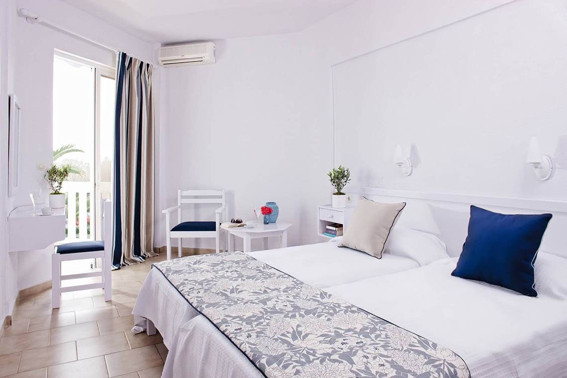 Schlafzimmer im Mrs Chryssana Beach Hotel mit zwei Betten, Kissen, Nachttischen und einem offenen Fenster.