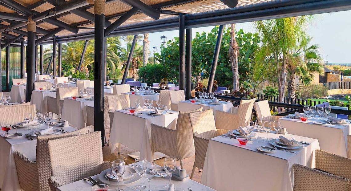 Außenbereich des Restaurants im Barcelo Fuerteventura Mar Hotel mit gedeckten Tischen und Stühlen, umgeben von Palmen und Blick auf den Garten.
