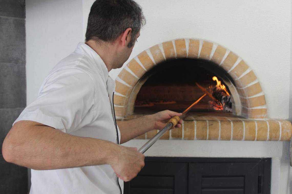 Ein Koch im Blue Aegean Hotel & Suites benutzt einen langen Holzstab, um Pizza im Ofen zu backen.
