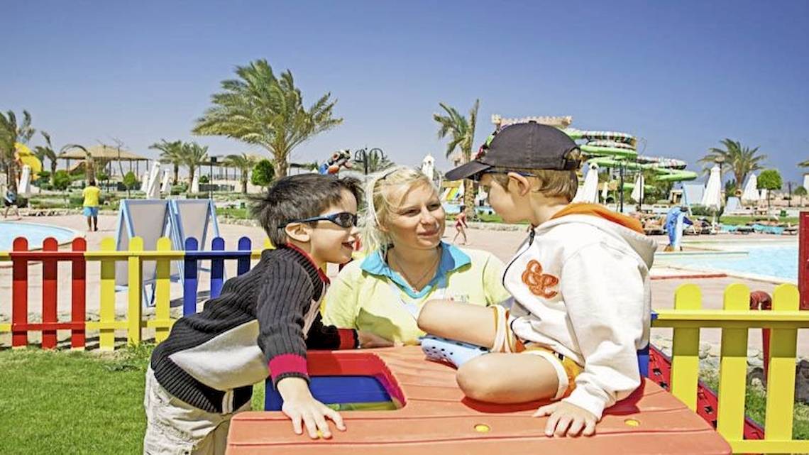Zwei Kinder und eine Frau spielen auf einem Spielplatz im Freien, mit einem Schwimmbad im Hintergrund.