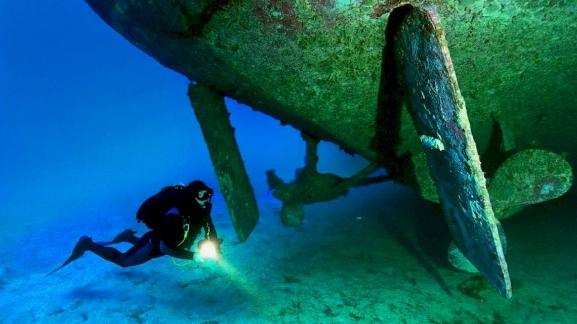 Taucher unter einem großen Schiffswrack mit Licht und Propellern.