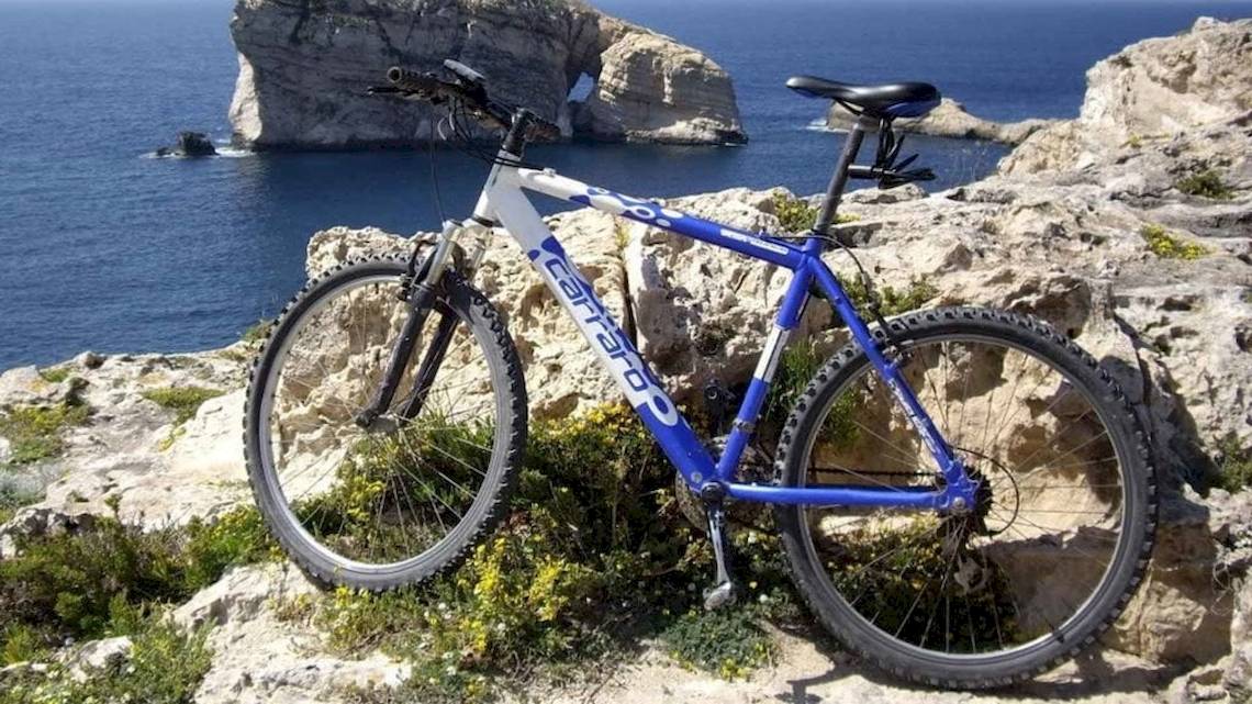 Blaues und weißes Mountainbike steht auf einem Felsen am Meer, mit Blick auf eine Felsformation.