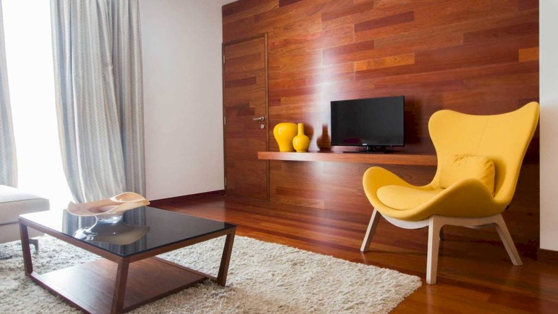 Ein Wohnzimmer mit einer holzverkleideten Wand, einem gelben Stuhl, einem Glastisch und einem Fernseher auf einem Regal.