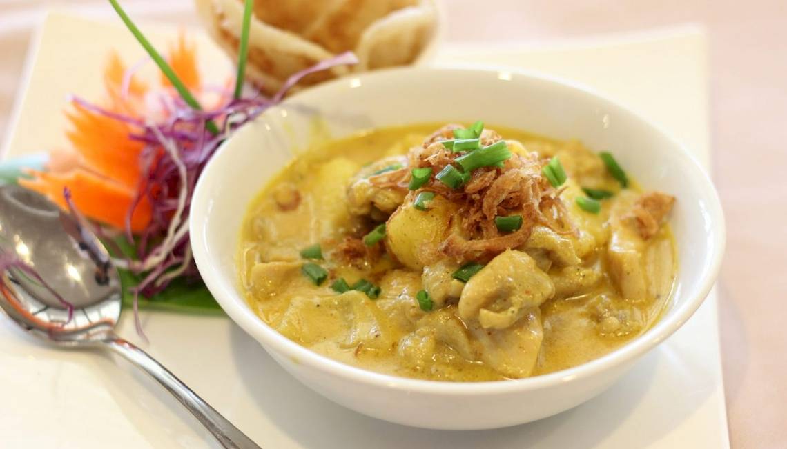 Ein weißer Bowl mit gelber Currysuppe, serviert mit einem Löffel und Beilagen, im Hotel Beyond Resort Krabi.