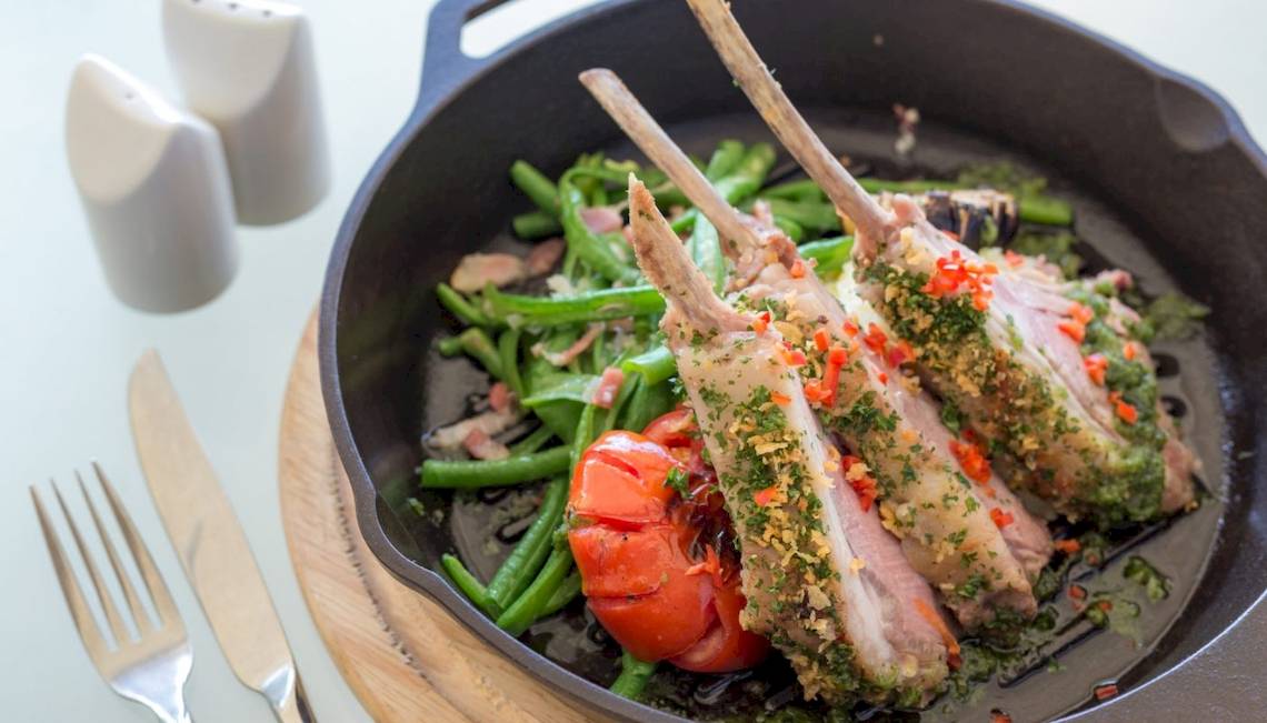 Ein Gericht in einer Pfanne mit gegrillten Lammkoteletts und Gemüse, einschließlich grüner Bohnen und Tomaten, serviert auf einem Holzuntersetzer mit Messer, Gabel und Salz- und Pfefferstreuer.