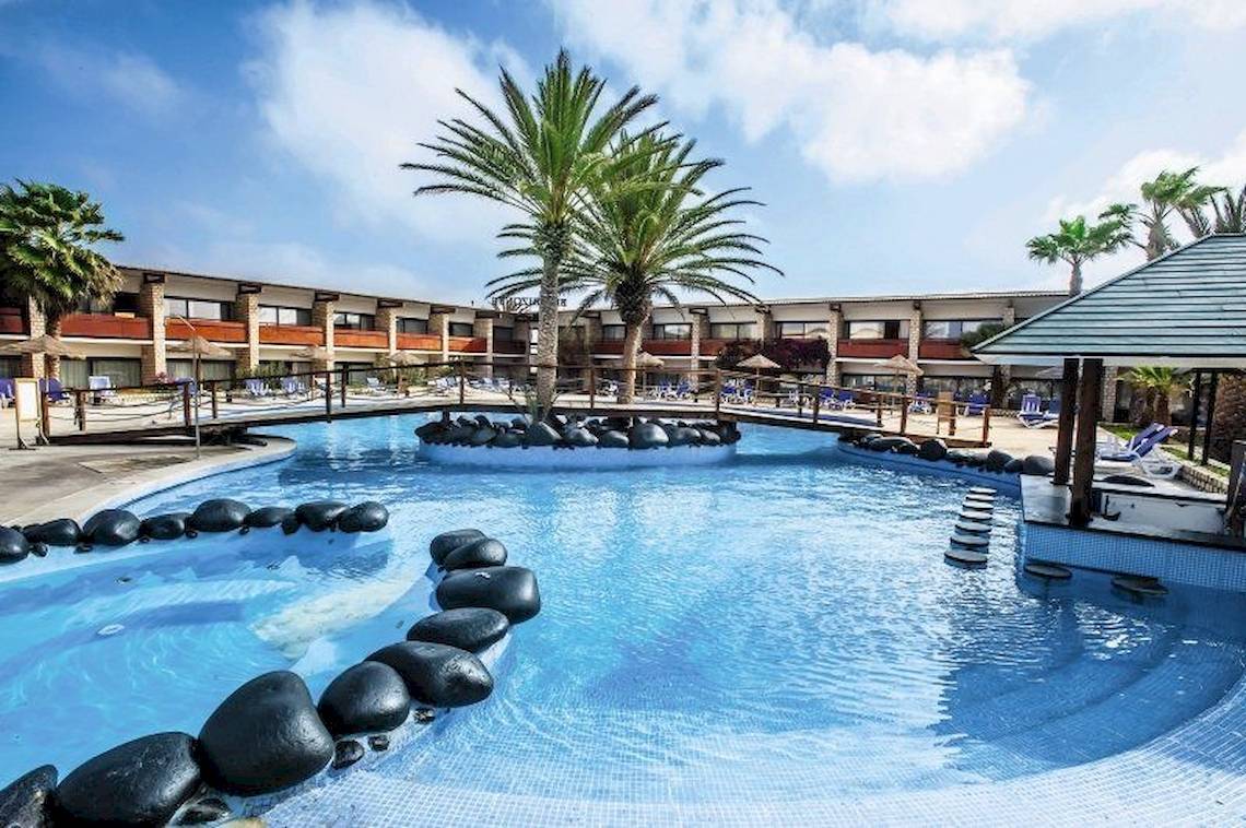 Außenpool des Oasis Belorizonte Hotels mit Palmen und Steinen, umrandet von Lounge-Bereichen.