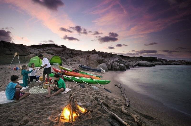 Familien-Campingplatz am Strand mit aufgestellten Kajaks, Zelten und einem brennenden Lagerfeuer