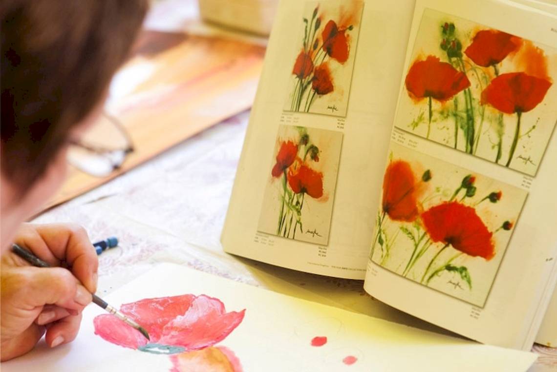 Eine Person malt eine rote Blume auf ein Blatt Papier mit einem Buch mit Blumenbildern im Hintergrund.