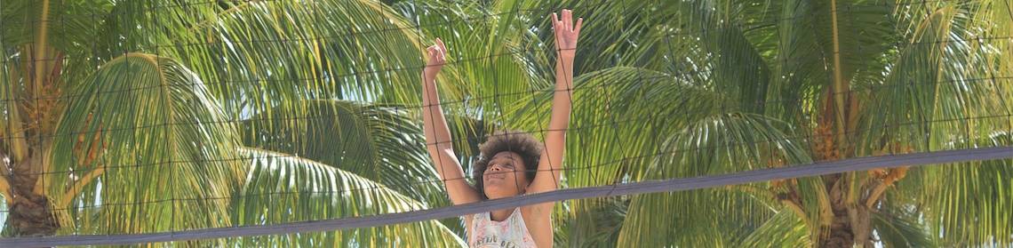 Eine Person spielt Beachvolleyball vor einem Netz und hohen Palmen.
