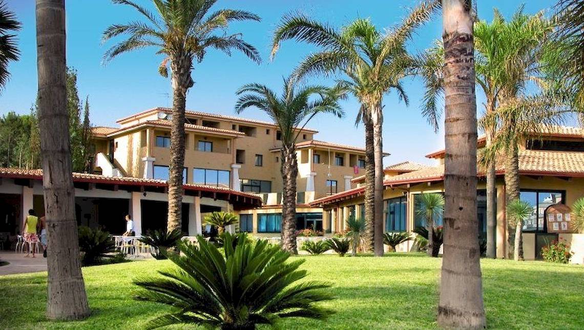 Außenansicht des TH Le Castella - Baia degli Dei Village Hotels mit Gebäuden, Palmen und grüner Rasenfläche.