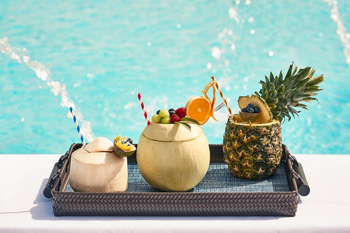 Cocktails in geschnittenen Kokosnuss- und Ananasschalen auf einem Tablett neben einem Pool im Raffles The Palm Dubai