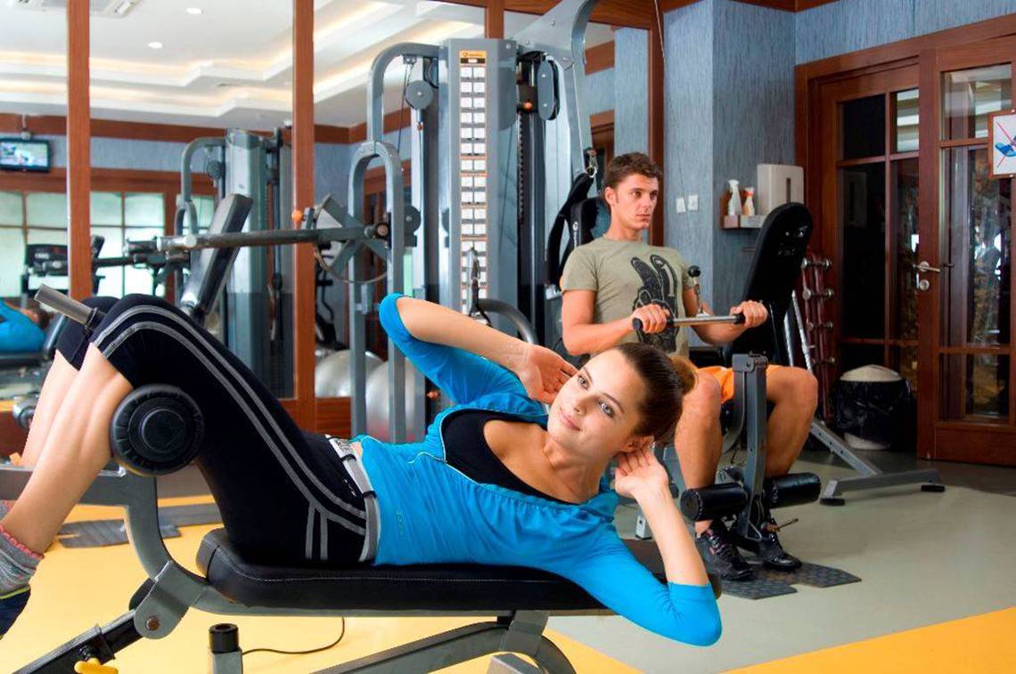 Fitnessstudio-Training mit Frau auf Bankpress-Maschine und Mann auf Langhantel-Bankdrücken.