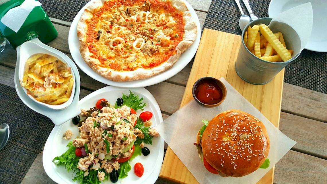 Verschiedene Teller mit Essen auf einem Holztisch, einschließlich einer Pizza, einem Salat, einem Burger und frittierten Pommes.